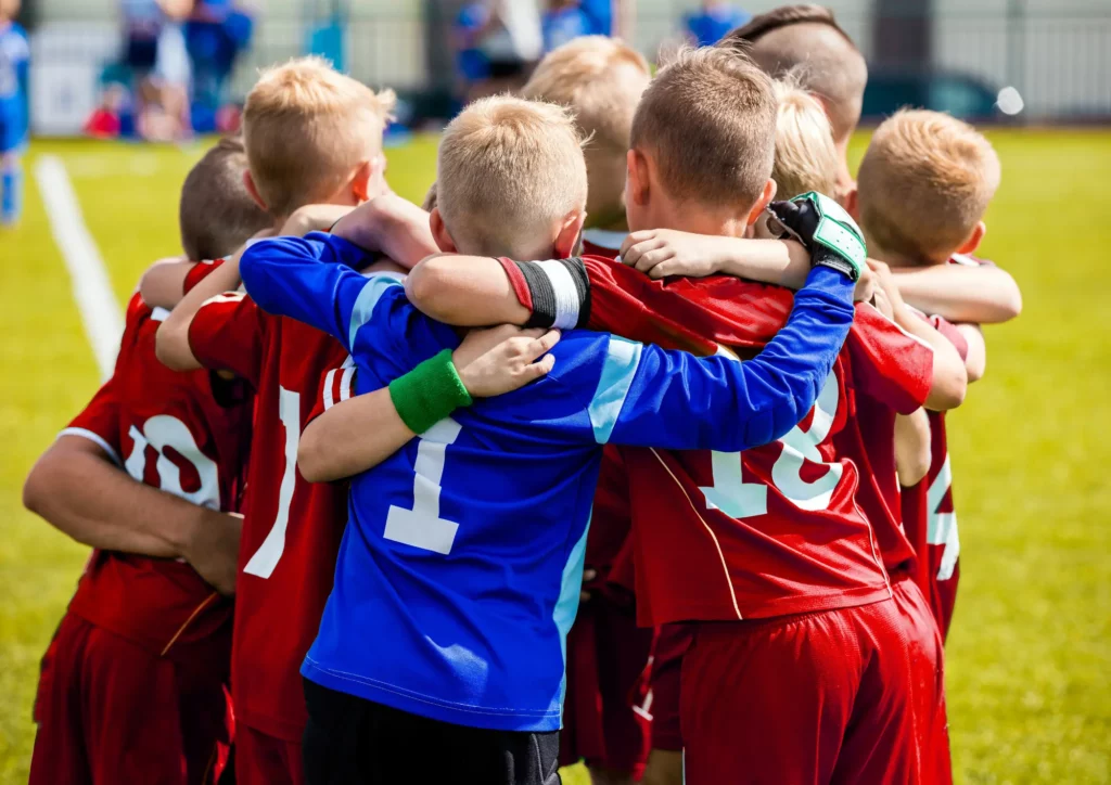 Youth soccer players in red jerseys form a tight huddle with a boy in blue at the center on a sunny grassy field.