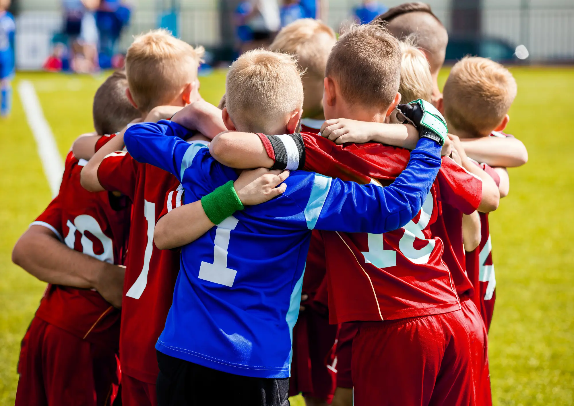 Youth soccer players in red jerseys form a tight huddle with a boy in blue at the center on a sunny grassy field.